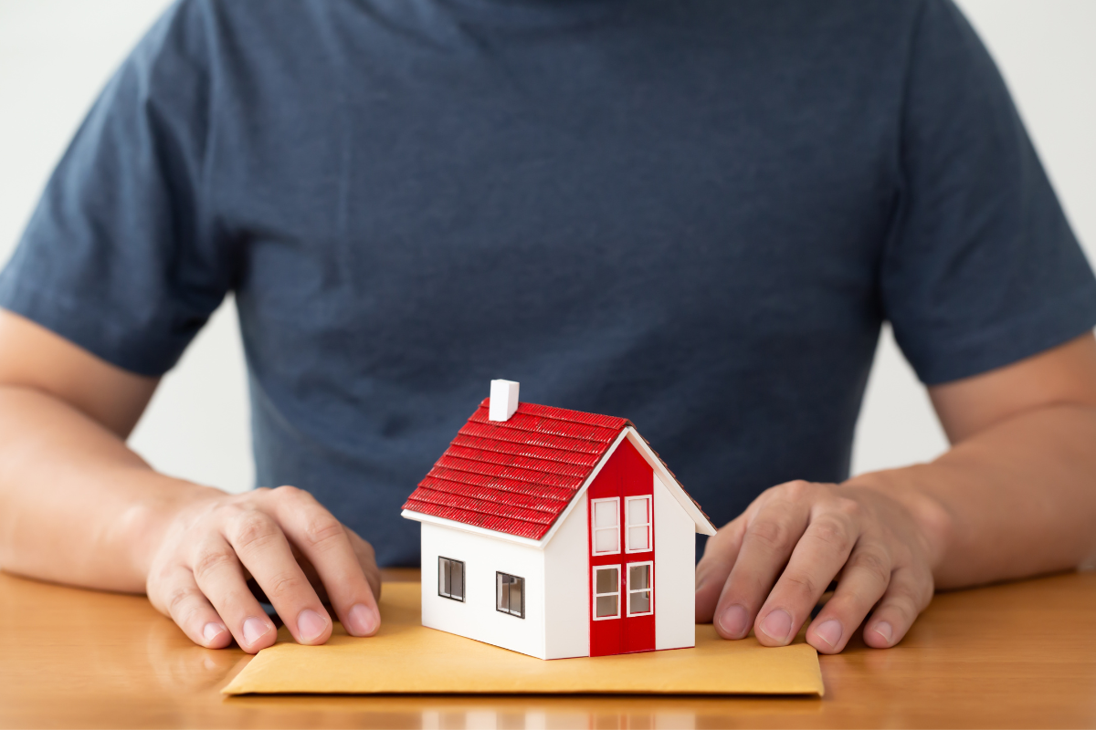 man in blue shirt sitting at wooden table with a manila envelope and a small red and white house sitting on top for benefits of refinancing