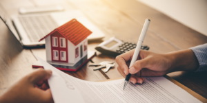 image of a person signing a document with a small house, calculator, keys and laptop in the background