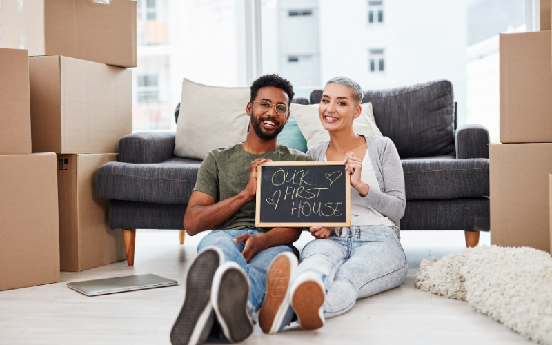 couple sitting on the floor holding a chalkboard sign that says Our First Home leaning against a gray couch for first home buyers guide 