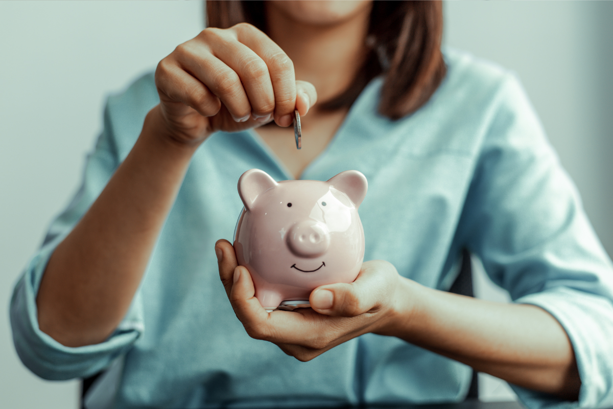 woman in blue shirt holding a pink piggy bank and dropping a coin into it for goal setting financial savings. 