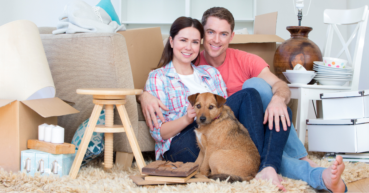 First time home buyers sitting on the floor in front of their couch with a small brown dog with them